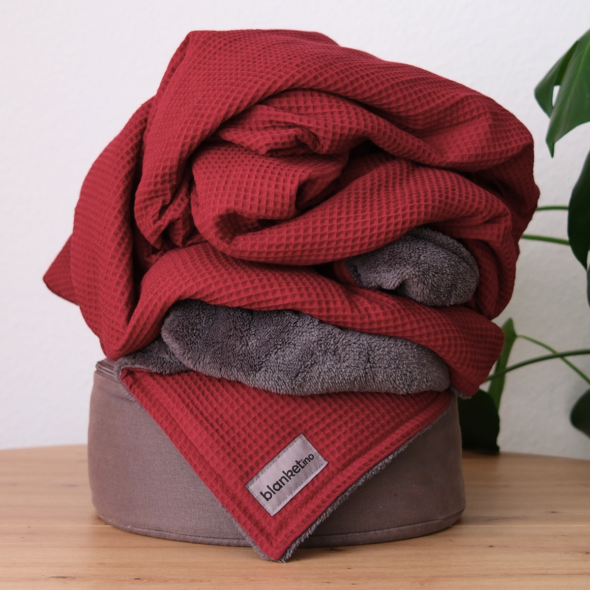 Weinrote und dunkelgraue Kuscheldecke "Vollkommen" von blanketino.de, locker auf einem Pouf drapiert, weiche Textur sichtbar