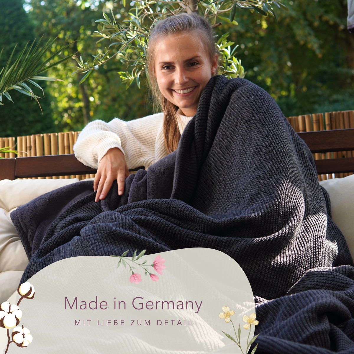 Dunkelgraue Kuscheldecke aus Baumwollstrick von blanketino.de, Frau sitzt entspannt auf Terrasse, hochwertig und bequem.