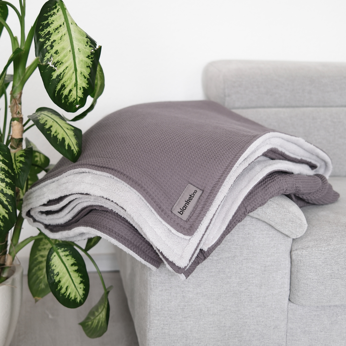 Gefaltete graue Kuscheldecke "Vollkommen" von blanketino.de auf einer Sofaarmlehne, weiches Textil, gemütliche Stimmung
