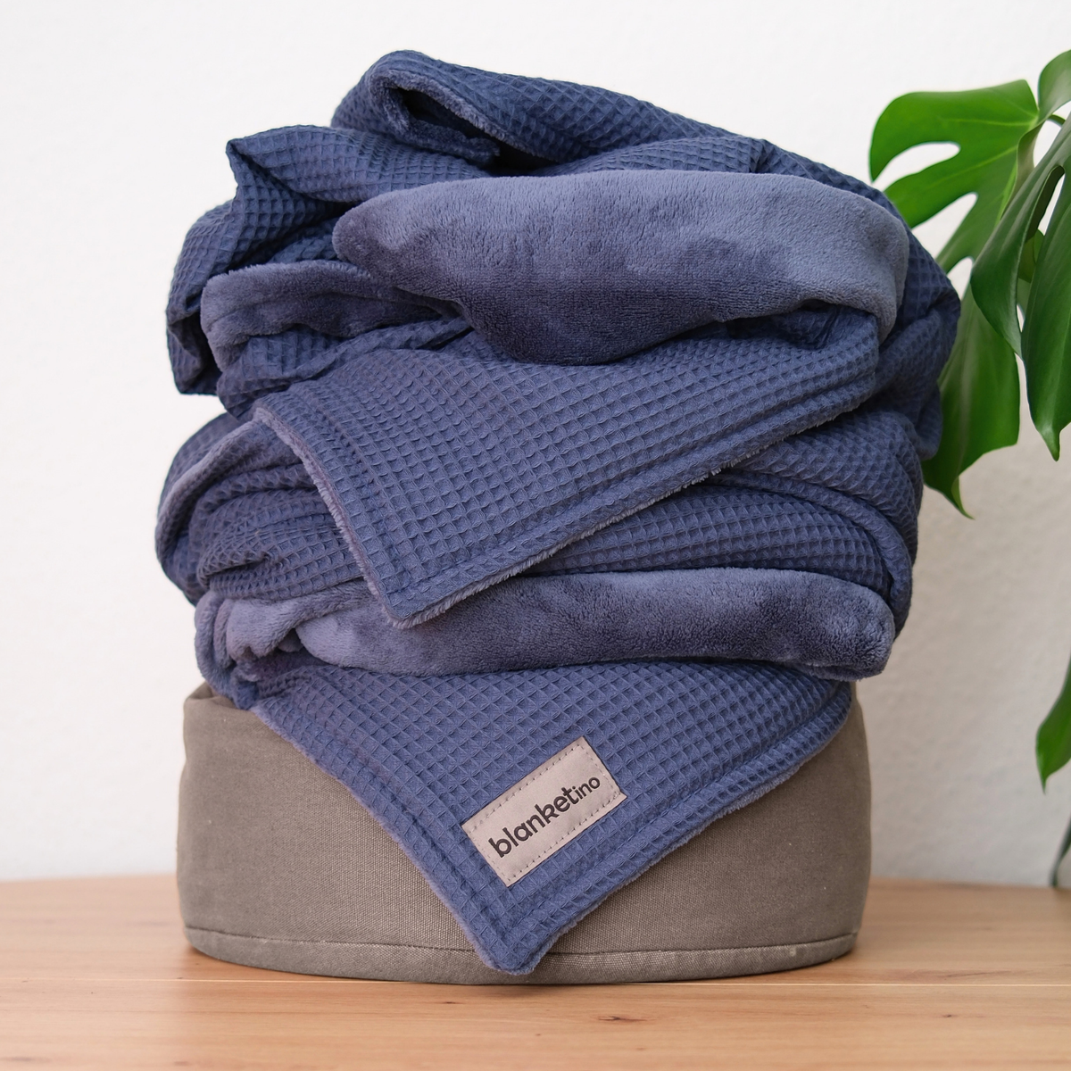 Dunkelblaue Kuscheldecke "Vollkommen" von blanketino.de, gefaltet auf einem Pouf, weiche Waffelpique-Struktur, gemütlich und einladend