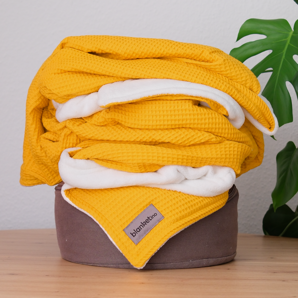 Weiche Kuscheldecke "Vollkommen" in Ocker Gelb und Sandweiß, gefaltet auf einem Pouf, mit Waffelpique-Struktur.